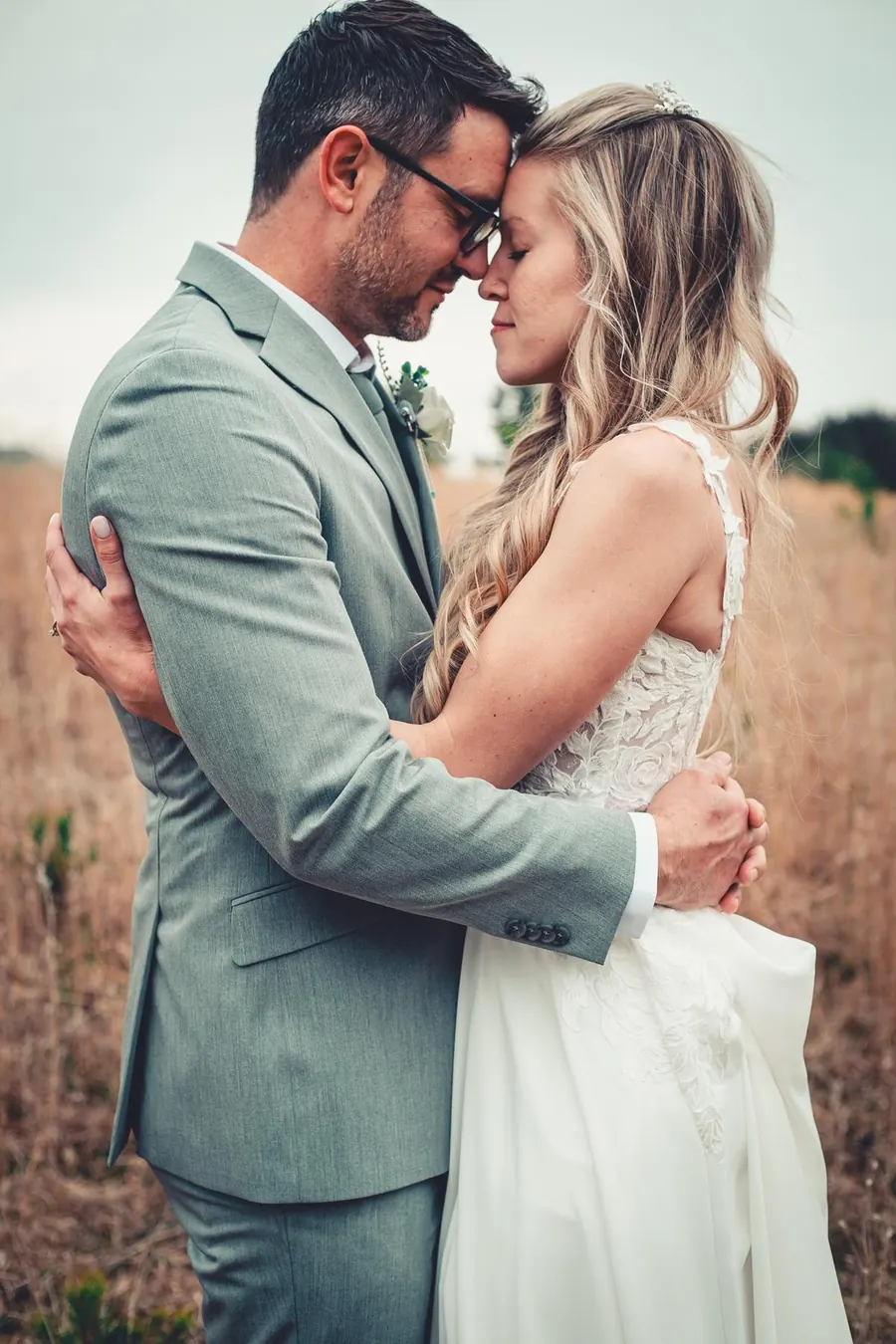 A bride and groom holding each other in a tall-grass field, foreheads together