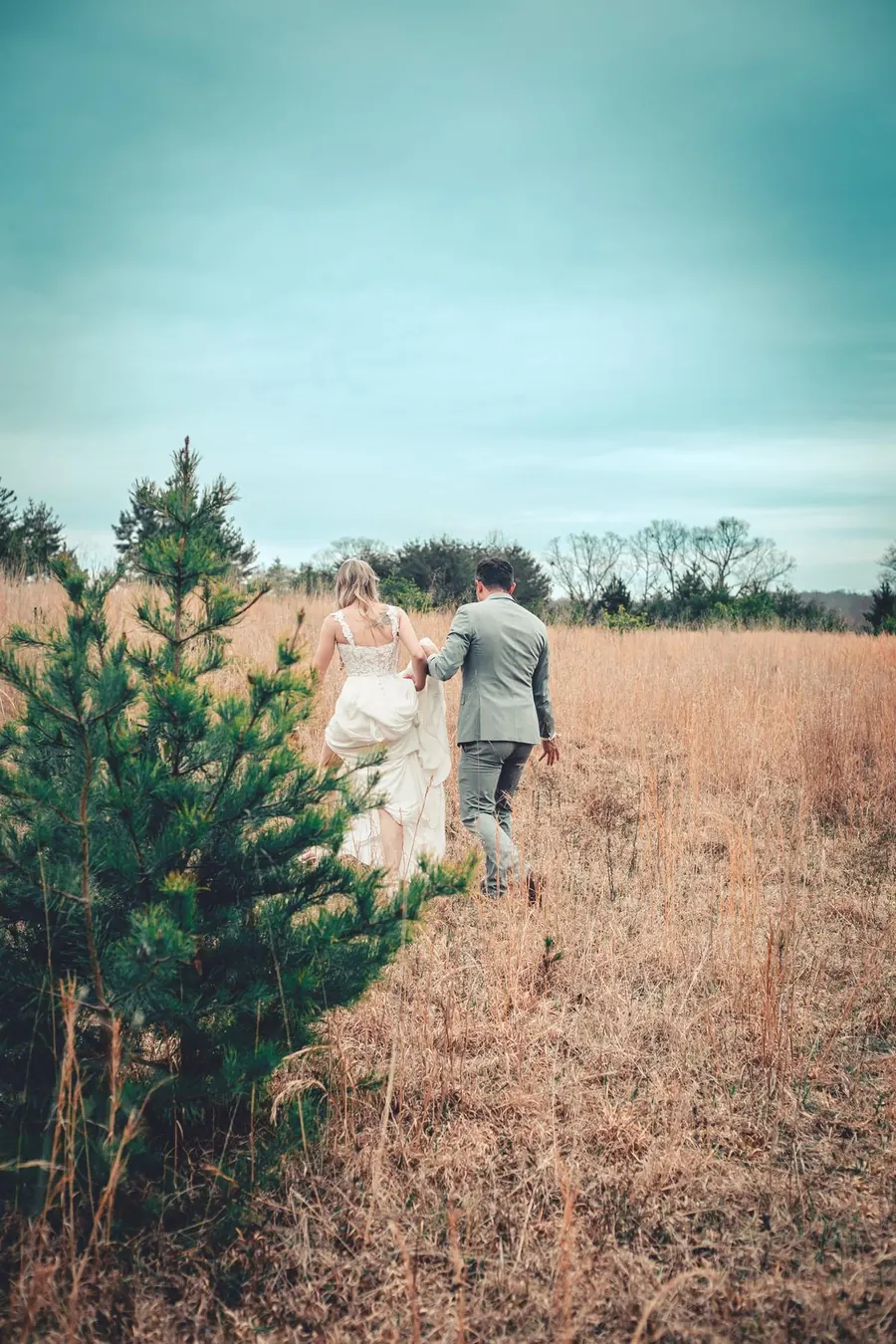 A bride and groom walking away through a wide autumn field under a soft blue sky
