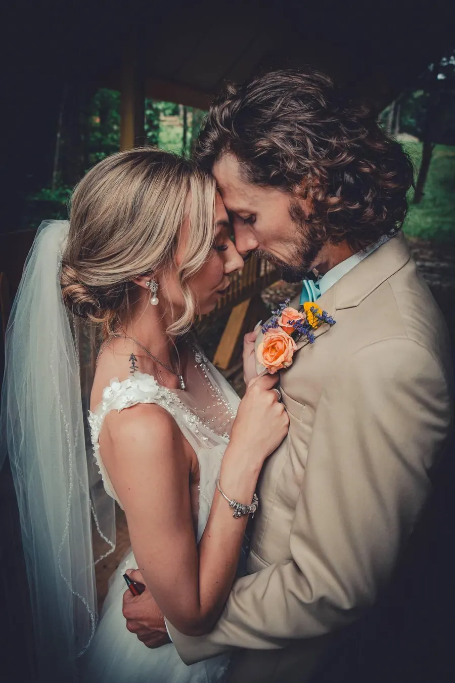 A bride in a long veil and a groom in a tan suit leaning in close, eyes closed