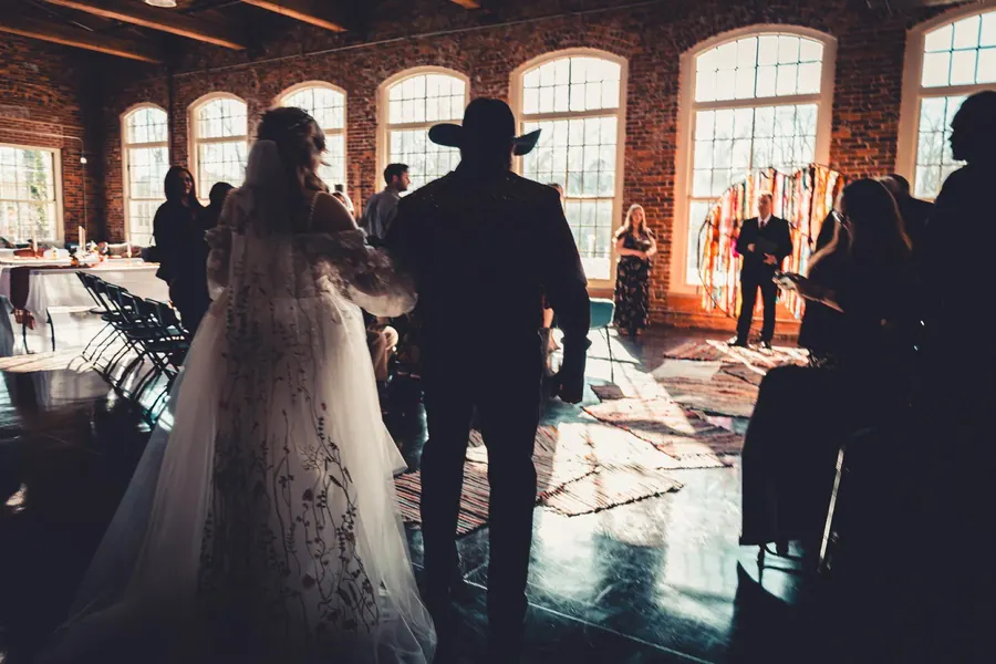 Guests in silhouette inside a brick hall with tall arched windows, the bride in the foreground