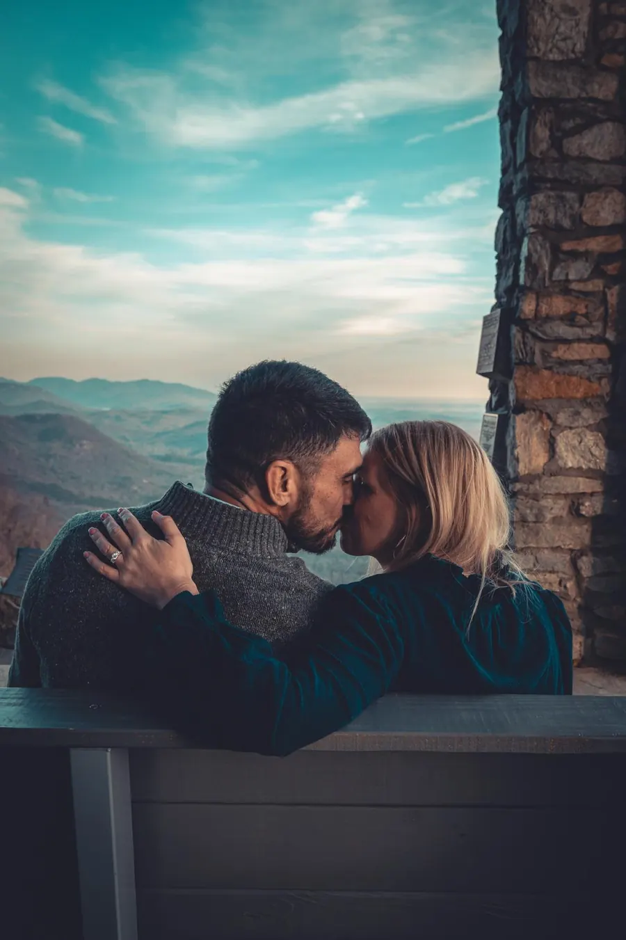 A couple on a bench at a stone lookout, mountains and open sky behind them