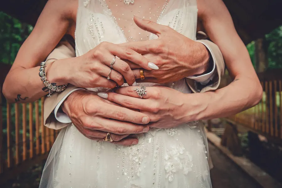 Close crop of a groom's hands wrapped around the bride's, rings showing against a beaded dress